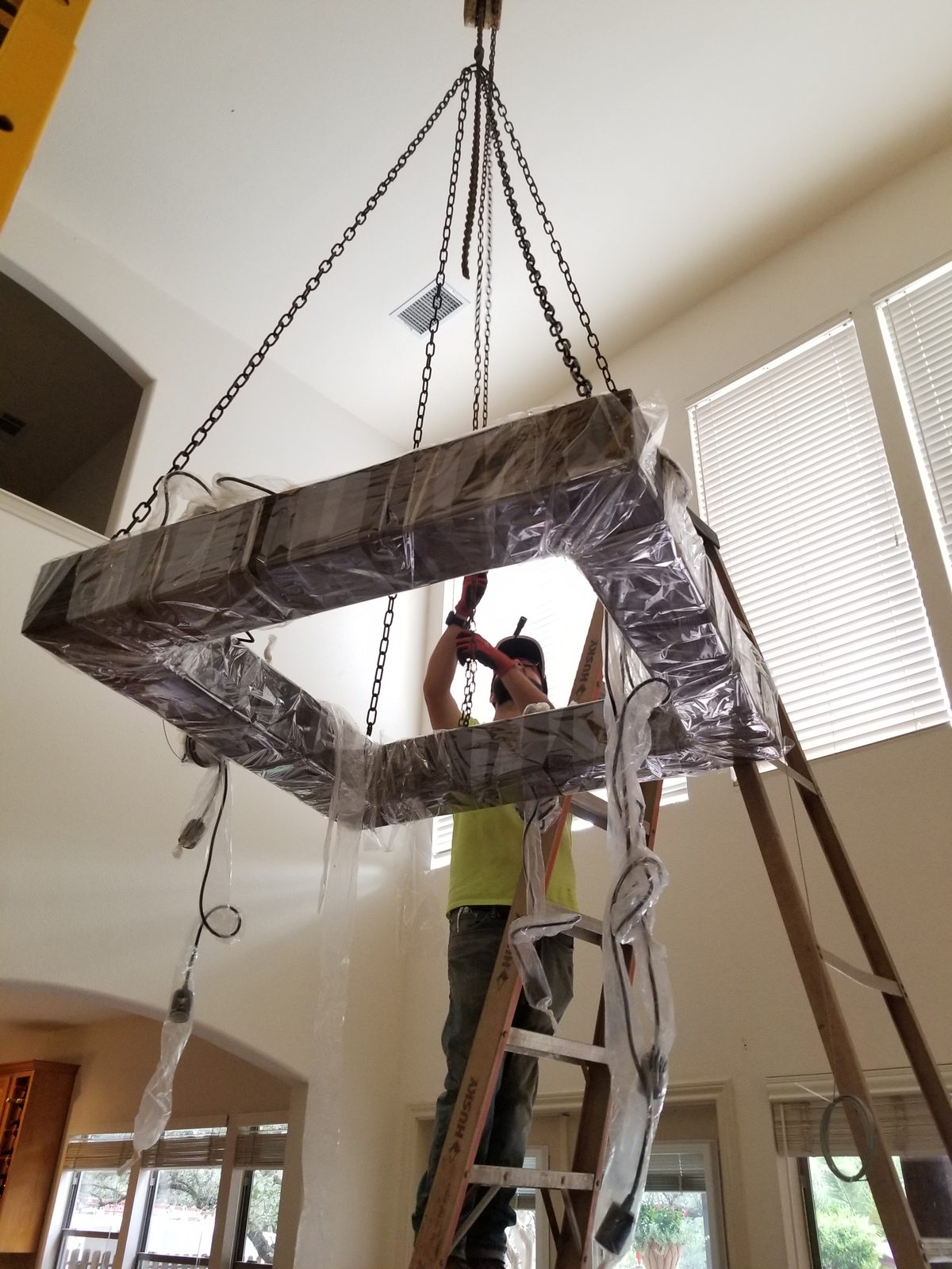 Electrician on a ladder installing a large chandelier still in protective wrap