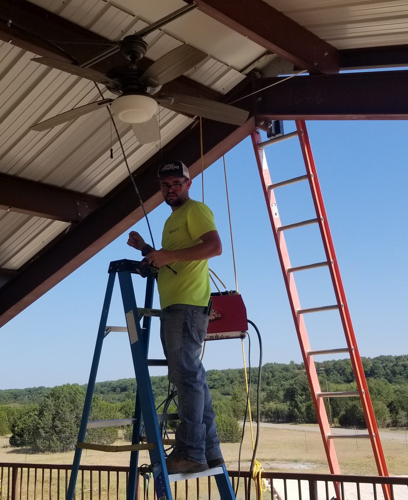 Electrician on a ladder installing a ceiling fan under an outdoor pavilion