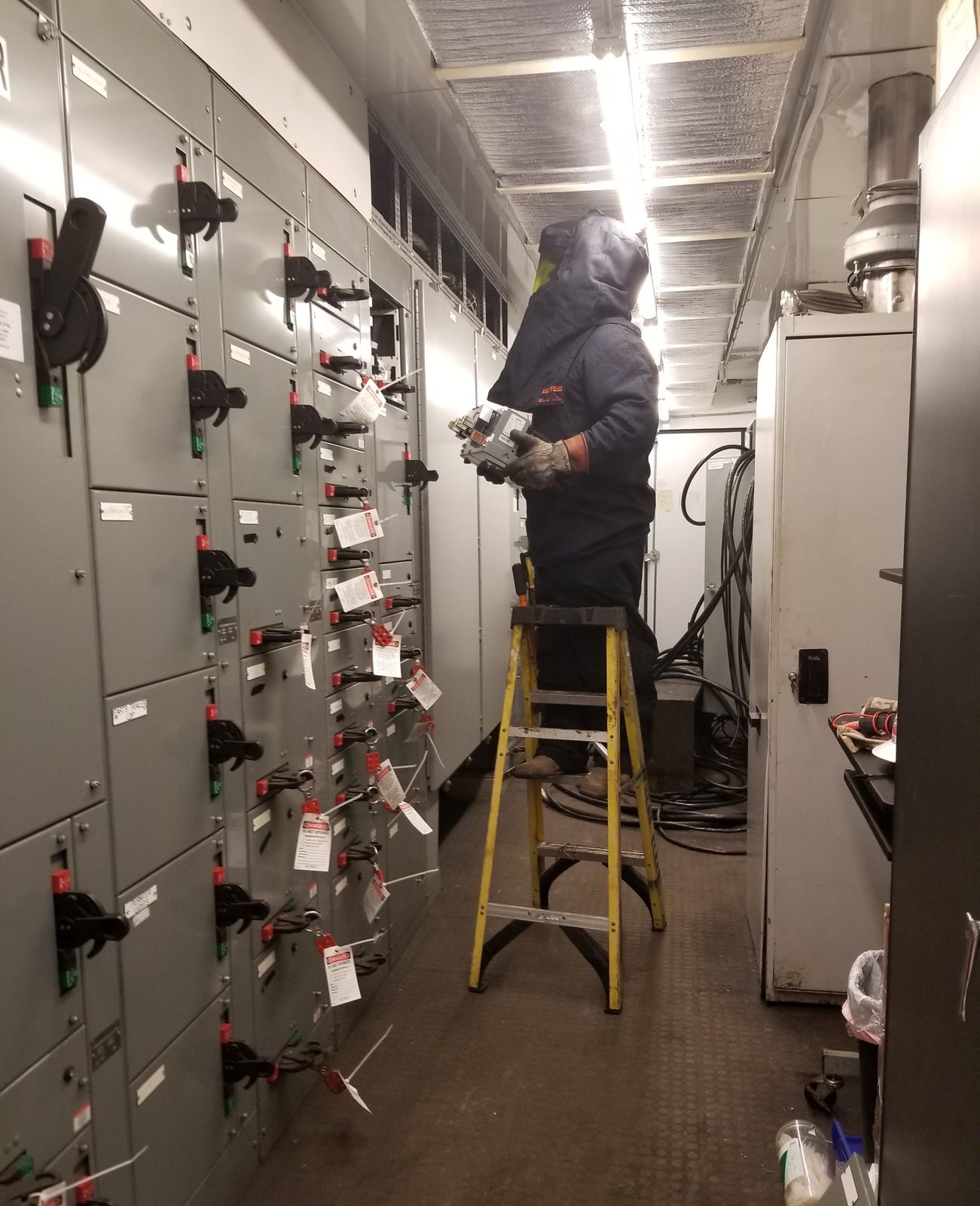 Electrician in an arc-flash hood on a ladder in an MCC aisle, lockout tags on the buckets